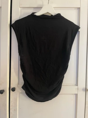 Svart ärmlös topp från Gina Tricot - Säljer en svart ärmlös topp från Gina Tricot i mjukt bomullsmaterial. Toppen har en snygg rynkad detalj längs sidan.