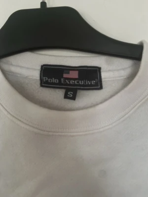 Vit sweatshirt från Polo Executive - Vit sweatshirt från Polo Executive med broderad logga och amerikansk flagga på bröstet. Klassisk rund halsringning och långa ärmar. Perfekt basic att ha i garderoben för en clean och enkel stil. ( fläckarna är från min kamera och finns inte på plagget ) 