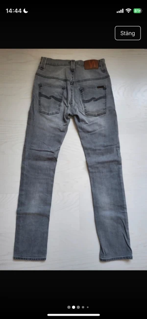 Grå Nudie Jeans, storlek 28/32 - Säljer ett par grå jeans från Nudie Jeans i storlek 28/32. Modellen har klassisk femficksdesign, raka ben och snygga detaljer på bakfickorna med Nudie-loggan. Jeansen är slim fitt 