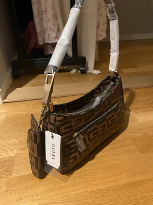 Brun axelväska från Guess med monogram - Snygg brun axelväska från Guess med stort G-mönster i svart och brunt. Väskan har guldfärgade detaljer, dragkedja upptill och en matchande liten plånbok på kedja. Tillverkad i syntetmaterial och har en bred axelrem.