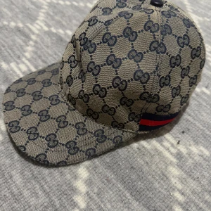 Gucci keps med GG-mönster och rödblå rand - Snygg keps från Gucci i beige med klassiskt GG-monogram i mörkblått över hela kepsen. Baksidan har en ikonisk röd och marinblå rand. Kepsen är tillverkad i bomullscanvas och har justerbar rem baktill för perfekt passform.
