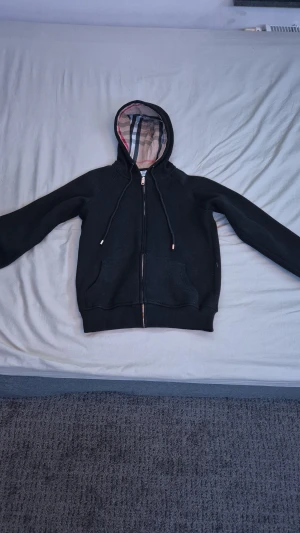 Svart Burberry hoodie med dragkedja - Svart hoodie från Burberry i storlek S med klassiskt rutigt foder i huvan. Tröjan har dragkedja framtill, snöre vid huvan och två fickor. Ingen logga på framsidan. Perfekt för dig som gillar stilrena och lyxiga detaljer.
