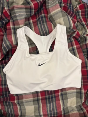 Vit sport-bh från Nike, storlek M - Säljer en vit sport-bh från Nike med klassisk svart swoosh-logga framtill. Modellen har brottarrygg och är tillverkad i ett mjukt, stretchigt syntetmaterial med meshdetaljer på insidan för extra ventilation. Perfekt för träning och aktiva dagar.