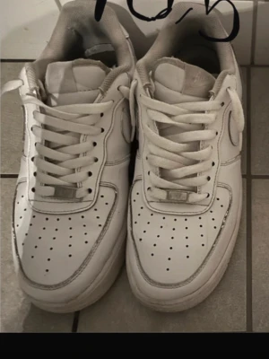 Nike Air Force 1 vita sneakers - Säljer ett par klassiska Nike Air Force 1 sneakers i helvitt skinn med perforerad tå och rund sula. Skorna har snörning och en stilren design som passar till många olika outfits. Perfekta för dig som gillar streetwear och vill ha en ikonisk sneaker i garderoben.