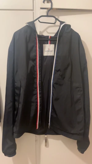 Moncler Windbreaker - Svart vindjacka från Moncler med huva och dragkedja i rött, vitt och blått. Jackan har lång ärm, Moncler-logga på ärmen och är tillverkad i lätt polyester. Perfekt för dig som vill ha en stilren och sportig look.                                                                NFC-tag finns.