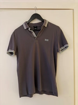 Grå pikétröja från Hugo Boss - Säljer en stilren grå pikétröja från Hugo Boss med klassisk krage och korta ärmar. Tröjan har vita och turkosa ränder på krage och ärmslut samt diskret Boss-logga på bröstet. Storlek M, är en regular fit 