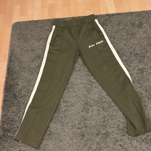 Palm Angels gröna mjukisbyxor med vita ränder - Snygga gröna mjukisbyxor från Palm Angels med vita sidoremsor och logga tryckt på framsidan. Byxorna har elastisk midja, raka ben och praktiska fickor. Perfekt för chill eller streetwear-stil.