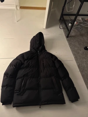 Svart pufferjacka från Zavetti Canada - Svart pufferjacka från Zavetti Canada med huva och snygga logopatchar på ärmen och insidan. Jackan har dragkedja framtill, två sidofickor med dragkedja och en loose passform. Perfekt för kalla dagar och har en riktigt modern vibe.