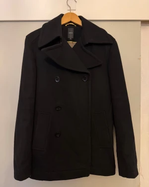 Svart skepparkavaj från Emilio - Svart skepparkavaj från Emilio med bred krage och dubbelknäppning. Jackan har två snedställda fickor fram och är fodrad med beige tyg och gul kant. Materialet är 70% ull och 30% polyamid, väldigt varm. Stilren och klassisk look. Small men passar även medium.