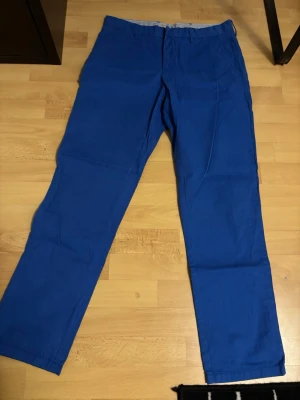 Blå chinos från Tommy Hilfiger - Snygga blå chinos från Tommy Hilfiger med raka ben och klassisk passform. Byxorna har två sidofickor fram och två bakfickor med knapp, samt bälteshällor. Materialet är mjuk bomull och färgen är intensiv blå. Perfekt för dig som gillar stilrena och bekväma byxor.