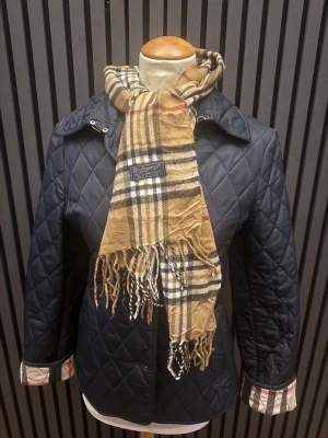 Scarf från Burberry - Säljer en quiltad Scarf från Burberry med klassisk snygga rutiga detaljer. Scarfen har en stilren look och passar perfekt till lager-på-lager. Ikonisk Burberry-vibe med rutmönster på detaljerna. SKICKAR INOM 24 TIMMAR 