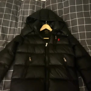 Ralph Lauren jacka puffer - Säljer en svart pufferjacka från Polo Ralph Lauren med huva och dragkedja framtill. Jackan har två sidofickor med dragkedja och en liten röd logga på bröstet. Perfekt för kalla dagar och riktigt snygg stil. Det ända är att det finns en knapp på armen om ja inte kan stänga, tror den är böjd men går nog att knäppa med en tång eller något.