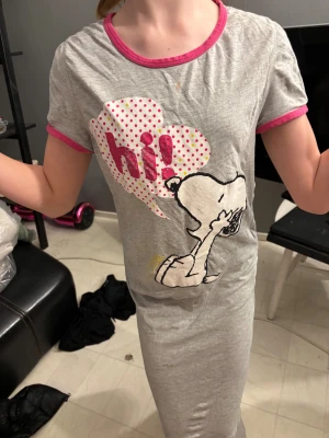 Grå Snoopy t-shirt med rosa detaljer - Säljer en grå t-shirt med tryck av Snoopy och pratbubbla med texten 'hi!' i rosa och prickigt mönster. T-shirten har korta ärmar och rosa kant vid hals och ärmslut. Perfekt för dig som gillar roliga prints och mjuka material.