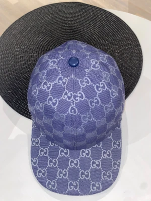 Blå keps från Gucci med GG-mönster - Snygg blå keps från Gucci med ikoniskt GG-mönster över hela kepsen. Justerbar rem bak i blått skinn och tillverkad i Italien. Kepsen har en klassisk form med böjd skärm och är gjord i bomullsmaterial med detaljer i skinn.