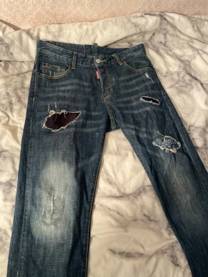 Dsquared2 slim jeans med slitningar - Säljer ett par blå Dsquared2 slim jeans i storlek 32 med coola slitningar, hål och färgstänk. Klassisk femficksmodell med läderpatch bak och snygga detaljer. Jeansen har en slim passform och är tillverkade i bomull. Perfekt för dig som gillar en edgy streetstil.