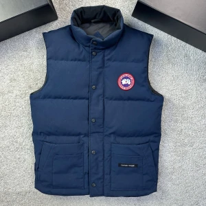Mörkblå dunväst från Canada Goose - Säljer en mörkblå dunväst från Canada Goose med hög krage och tryckknappar framtill. Västen har två stora fickor och en klassisk logotyp-patch på bröstet. Perfekt för kyliga dagar när du vill hålla stilen och värmen. Moselle heter Freestyle Gilet och är i storlek S. 