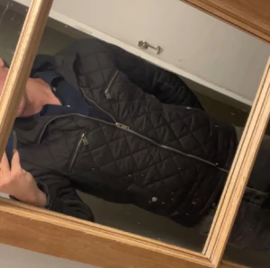 Svart quiltad jacka med dragkedjor - Säljer en svart quiltad jacka med snygga dragkedjor på bröstet och framtill. Jackan har hög krage och långärmade ärmar, perfekt för dig som vill ha en stilren look. Passformen är normal och materialet känns som polyester. Riktigt schysst jacka för dig som gillar klassisk stil.