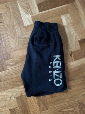Svarta Kenzo shorts med tryck - Svarta shorts från Kenzo med stort vitt tryck på ena benet. Modellen har elastisk midja med snörning, två sidofickor och två bakfickor. Mjukt bomullsmaterial som är skönt mot huden och passar perfekt till chill eller häng.
