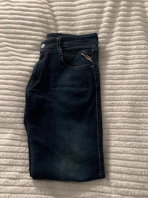 Replay Anbass mörkblå jeans - Snygga Replay Anbass jeans i mörkblå tvätt med klassisk femficksdesign och smal passform. Jeansen har diskreta slitningar och detaljer som Replay-logga på fickan. Perfekta för dig som gillar stilrena och moderna jeans med lite stretch.