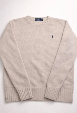 Beige stickad tröja Polo Ralph Lauren - Snygg beige stickad tröja från Polo Ralph Lauren med klassisk rund hals och ribbade muddar. Tröjan har en mörkblå broderad logga på bröstet och är långärmad. Perfekt för lager-på-lager under höst och vinter.