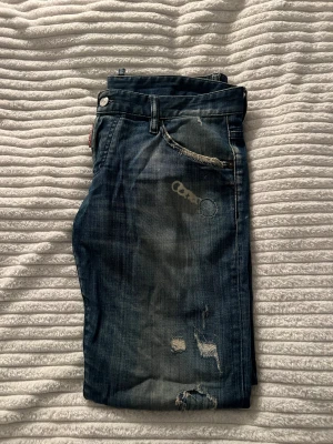 DSQUARED 2 Jeans med slitningar - Säljer ett par DSQUARED 2 jeans i blå tvätt med coola slitningar och trasiga detaljer på både fram- och baksida. Jeansen har en rak passform och är gjorda i jeansmaterial.