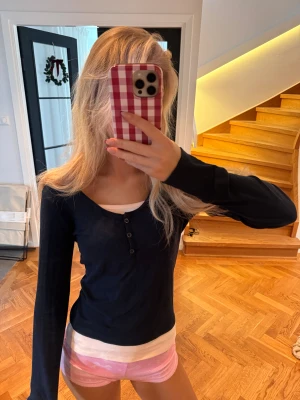 Svart långärmad topp med knappar - Säljer en mörkblå och ljusrosa långärmad topp med rundad hals och tre knappar framtill. Modellen är slim och sitter snyggt mot kroppen. Materialet känns mjukt och stretchigt, troligen bomull eller polyester. Perfekt att styla med jeans eller kjol.