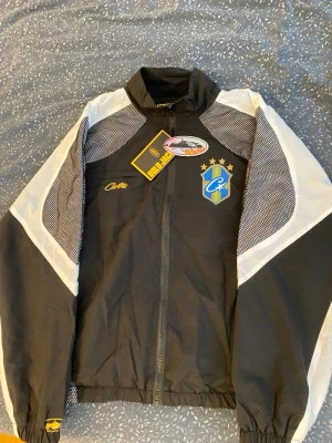 Svart och vit vindjacka från Corteiz - Snygg vindjacka från Corteiz i svart och vitt med coolt broderat emblem på bröstet och gul logga. Jackan har dragkedja framtill, långärmade armar och detaljer i meshmönster på axlar och ärmar. Priset är inte ristat i sten och går att diskuteras