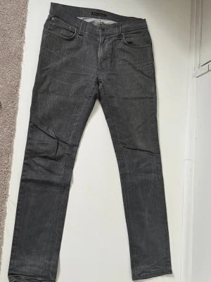 Grå jeans från Nudie Jeans - Säljer ett par grå jeans från Nudie Jeans med diskret strimmigt mönster. Klassisk femficksmodell med raka ben och normal passform. Materialet är jeans och färgen är mörkgrå, vilket ger en stilren vibe. Perfekt för dig som gillar enkel och clean stil.w32 L34