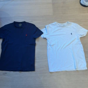 2-pack Polo Ralph Lauren t-shirts - Två klassiska t-shirts från Polo Ralph Lauren, en i marinblått med röd broderad logga och en i vitt med mörkblå logga. Båda är kortärmade och har en normal passform. Perfekta till jeans eller shorts för en clean och stilren look. med ett litet hål oå den blåa, synns knappt. Ena tröjan är barn men passar Xs
