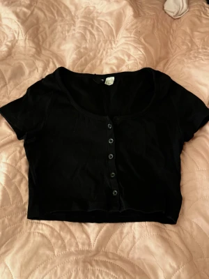 Svart croppad topp med knappar - Säljer en svart croppad topp från h&m med korta ärmar och rund halsringning. Toppen har en rad svarta knappar framtill och är gjord i mjuk bomull. 