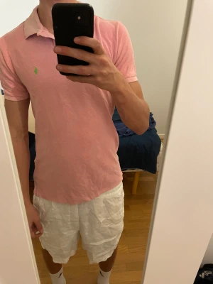 Polo Ralph Lauren piké - Säljer en klassisk rosa pikétröja från Polo Ralph Lauren med korta ärmar och krage. Tröjan har en grön broderad logga på bröstet och knappar vid halsen. Materialet är mjuk bomull och passformen är normal. Perfekt för sommarens chill dagar.