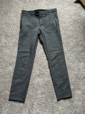 Grå kostymbyxor från J.Lindeberg - Snygga grå kostymbyxor från J.Lindeberg i storlek 48. Byxorna har ett klassiskt, rakt snitt med bälteshällor, framfickor och bakfickor med knapp. Materialet är mjukt och har en diskret struktur som ger en stilren look.