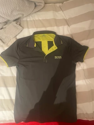 Svart t-shirt från Hugo Boss - Säljer en svart t-shirt från Hugo Boss med gul insida på kragen och gul logga på bröstet. T-shirten har korta ärmar och en klassisk krage med knappslå. Perfekt för dig som gillar stilrena märkesplagg med en färgklick.