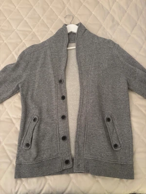 Grå kofta med knappar - Säljer en stilren grå kofta med knappar framtill och två fickor med knappdetaljer. Koftan har lång ärm och en hög krage som ger en chill vibe. Perfekt att slänga på sig när det är lite kyligt ute. Använd fåtal gånger