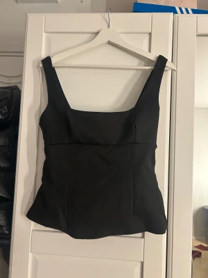 Svart linnetopp med rosett på ryggen  - Säljer en svart linnetopp med breda axelband och fyrkantig halsringning. Toppen har en stor dekorativ rosett på ryggen som ger en extra cool touch. Materialet är slätt och har en lätt glansig finish. Passformen är normal och toppen är ärmlös. Dragkedja på sidan och ganska stor i storleken 