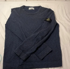 Blå långärmad tröja från Stone Island - Säljer en mörkblå långärmad tröja från Stone Island med klassisk rund hals och ikonisk patch på vänster ärm. Tröjan har en normal passform och är tillverkad i mjukt bomullsmaterial som känns skönt mot huden. Perfekt för dig som gillar stilrena och enkla plagg.