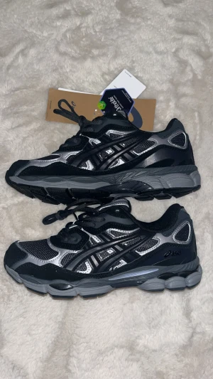 Svarta och silver Asics sneakers - Säljer ett par snygga Asics sneakers i svart och silver med meshpaneler och detaljer i syntet. Skorna har rund tå, platt sula och snörning framtill. Perfekta för dig som gillar sportig stil och vill ha bekväma skor med cool design.