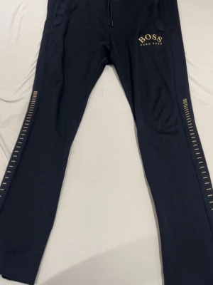Svarta mjukisbyxor från Hugo Boss M - Svarta mjukisbyxor från Hugo Boss med guldfärgade detaljer och logga på benet. Byxorna har snörning i midjan och guldfärgad text på insidan av linningen. Snygga ränder längs benen ger extra stil. Perfekta för chill eller streetwear.