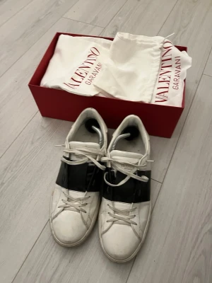 Valentino Garavani Open sneakers vit/svart - Säljer ett par Valentino Garavani Open sneakers i vitt skinn med bred svart rem över mitten. Klassisk låg modell med snörning och rund tå. Snygga detaljer med nitar på hälen och platt sula. Kommer med originalkartong och dustbags.