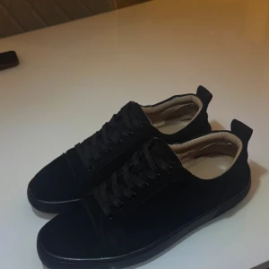 Loubitin skor - Säljer ett par svarta sneakers från Christian Louboutin med klassisk röd sula och snygg snörning. Skorna har rund tå och är tillverkade i mocka med mjukt beige foder. Perfekta för dig som vill ha en lyxig och stilren look.