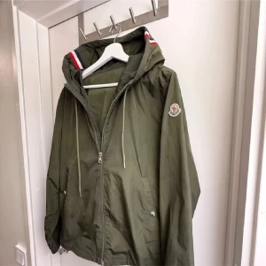 Grön vindjacka från Moncler - Snygg grön vindjacka från Moncler med huva och dragkedja framtill. Jackan har vita och röda detaljer på huvan, samt Moncler-logga på ärmen. Perfekt för blåsiga dagar och har praktiska fickor med knappstängning.