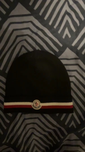 Svart Moncler mössa med röd/vit rand - Snygg svart mössa från Moncler med klassisk röd och vit rand längst ner samt Moncler-logga framtill. Mössan är ribbstickad och har en clean, stilren look som passar perfekt till vintern. Tillverkad i mjuk ull för maximal komfort. Finns även kvitto. Köptes nu i år men tröttnat på den då jag inte gillar passformen. 