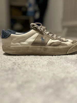 Polo Ralph Lauren sneakers i beige och blå - Snygga sneakers från Polo Ralph Lauren i beige och vitt skinn med mockadetaljer och blå textil på hälen. Klassisk snörning och rund tå, med Polo-logga broderad på sidan och bak. Perfekt för dig som gillar stilrena och sportiga skor.