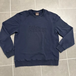 Mörkblå sweatshirt från Oscar Jacobson L - Snygg mörkblå sweatshirt från Oscar Jacobson med broderad logga framtill. Tröjan har rund hals, ribbade muddar vid ärmslut och nederkant samt långärmad passform. Perfekt för dig som gillar stilrena och klassiska plagg.