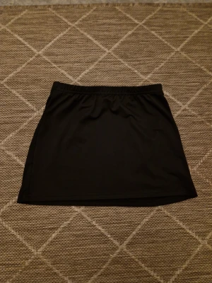 Svart kortkjol  - En enkel och stilren svart kortkjol. Kjolen har en rak passform och är tillverkad i ett mjukt syntetmaterial som känns bekvämt mot huden. Perfekt att matcha med en cool t-shirt eller hoodie för en avslappnad look.