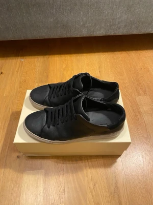 Svarta sneakers från Axel Arigato - Snygga svarta sneakers från Axel Arigato med klassisk rund tå och platt vit sula. Skorna har svart snörning och är tillverkade i slätt skinn som ger en stilren vibe. Perfekta för dig som gillar minimalistisk design och vill ha ett par schyssta sneakers i garderoben. Priset kan diskuteras!