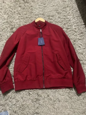 Mörkröd bomberjacka från GANT - Snygg mörkröd bomberjacka från GANT med klassisk ståkrage och dragkedja framtill. Jackan har två snedställda fickor och ribbade muddar vid ärmslut och nederkant. Materialet är polyester och jackan har en stilren look med diskret GANT-logga på bröstet.