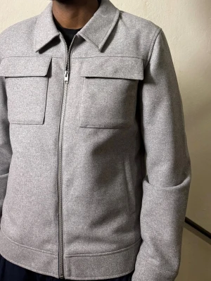 Grå overshirt med dragkedja - Snygg grå overshirt med rak passform och stilrena detaljer. Jackan har två stora bröstfickor med lock, klassisk krage och hel dragkedja framtill. Materialet är mjukt och känns som ull, perfekt för lager-på-lager. Passar dig som gillar minimalistisk stil.