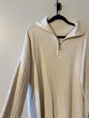 Mysig beige ribbad tröja med zip - Säljer en beige långärmad tröja med ribbad struktur och zip framtill. Tröjan har en hög krage och är tillverkad i ett mjukt stickat material. Passformen är loose och den neutrala färgen gör den enkel att matcha. Helt ny, endast testad 