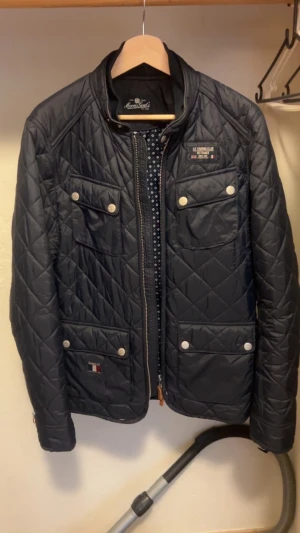 Mörkblå quiltad jacka från Morris - Quiltad mörkblå jacka från Morris med fyra framfickor och silverfärgade knappar. Jackan har ståkrage, dragkedja och snygga detaljer som patch på bröstet. Perfekt för dig som gillar stilren och sportig look. Jackan är M men kan passa S